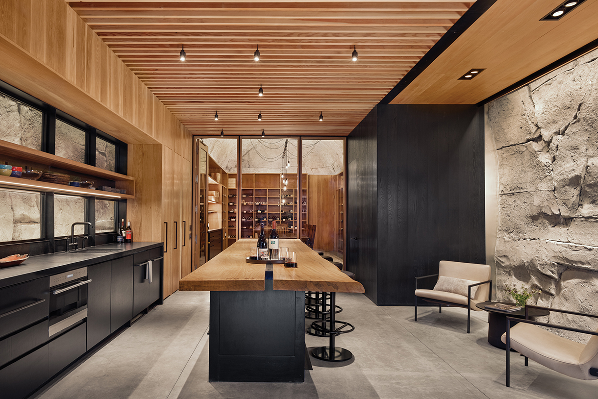 A spacious and modern, raw white oak kitchen with concrete walls sculpted to look like natural limestone, beamed ceilings, a long wooden cedar table, and a wine cellar stocked with bottles of wine at the back of the kitchen.